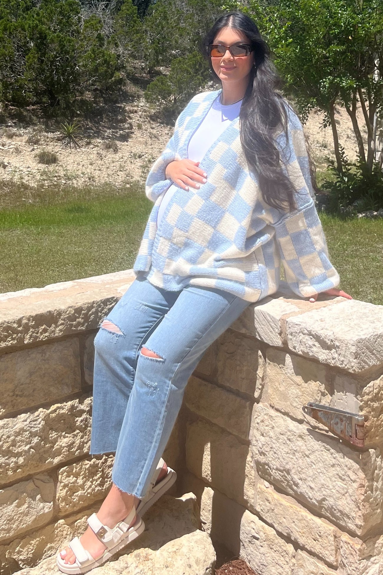 woman wearing a light blue and white checkered maternity cardigan, white top, and distressed jeans, holding her baby bump