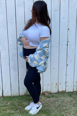 woman wearing a white short sleeve top and black leggings, holding her baby bump, layered with a blue plaid oversized shirt