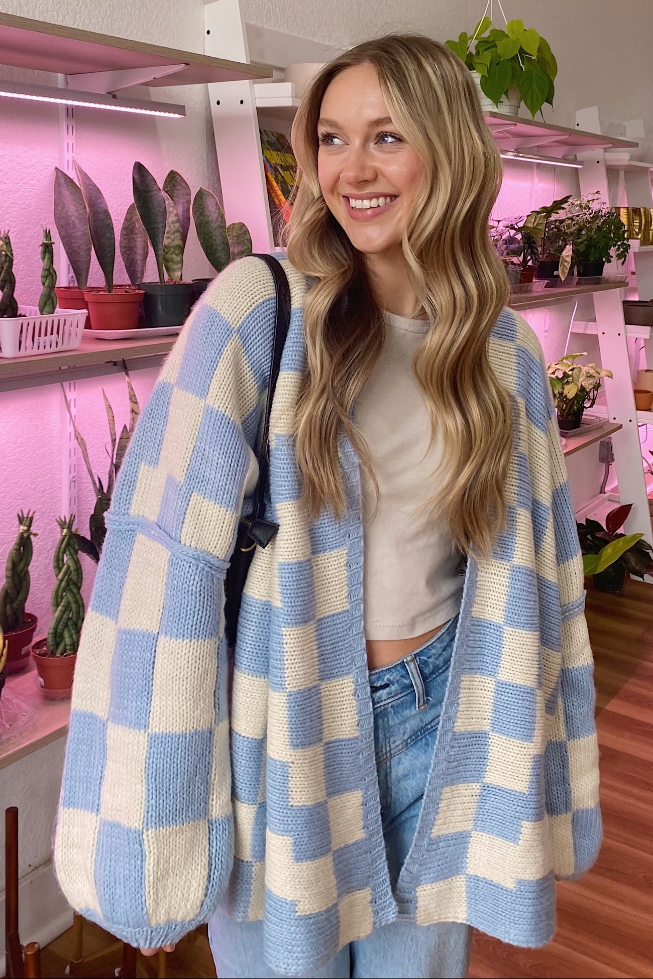 woman wearing a blue and cream checked cardigan over a white top, smiling in a plant-filled room