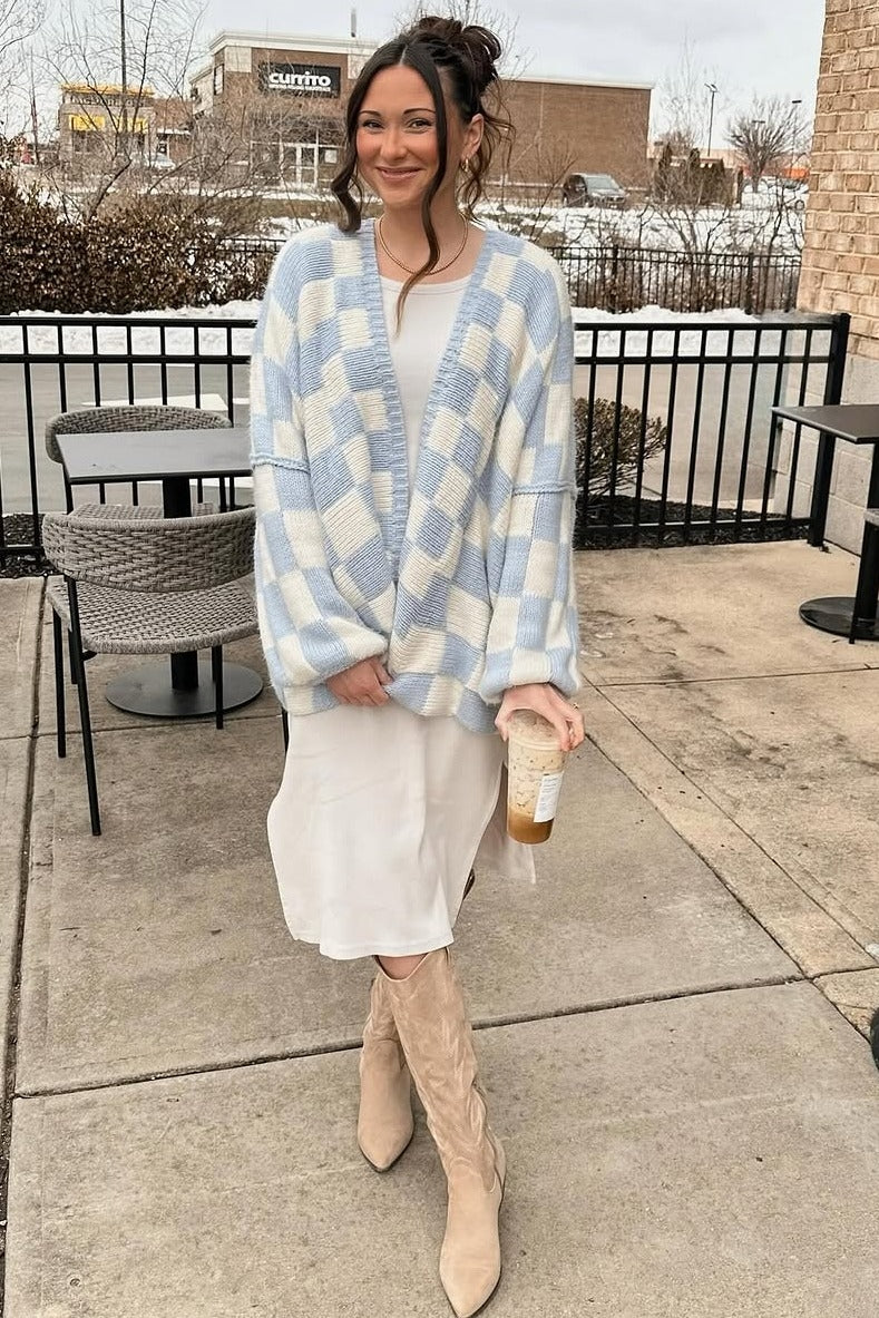 woman wearing a light blue and white checkered oversized cardigan, beige knee-high boots, and a white dress while holding a drink
