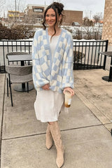 woman wearing a light blue and white checkered oversized cardigan, beige knee-high boots, and a white dress while holding a drink