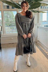 woman wearing a black and white gingham long sleeve dress with tiered hem and knee-high white boots, standing indoors