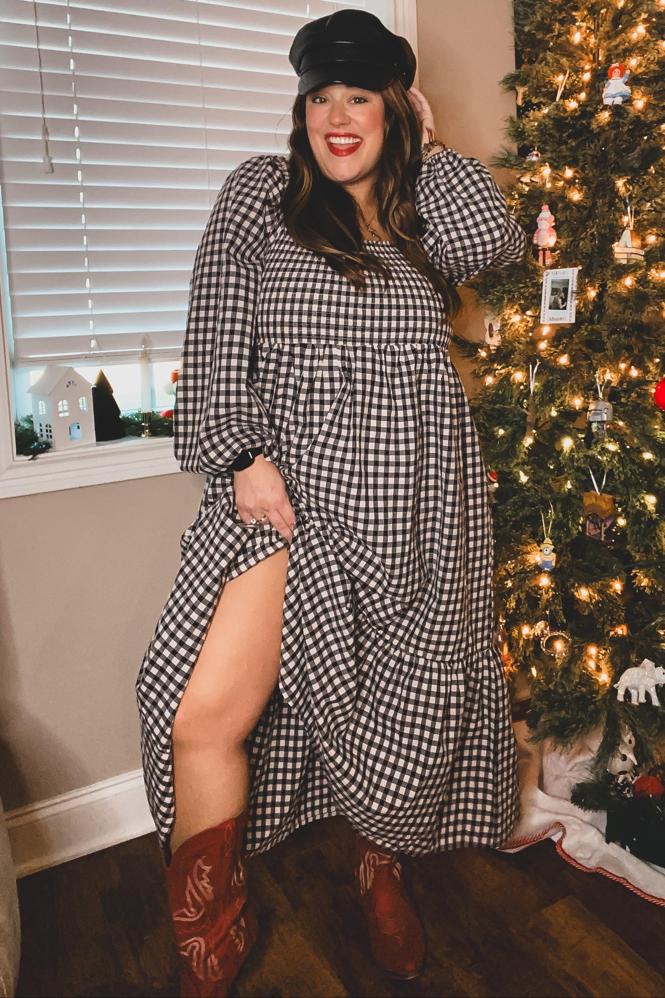 woman wearing a black and white gingham long sleeve maxi dress and brown cowboy boots, posing beside a decorated Christmas tree