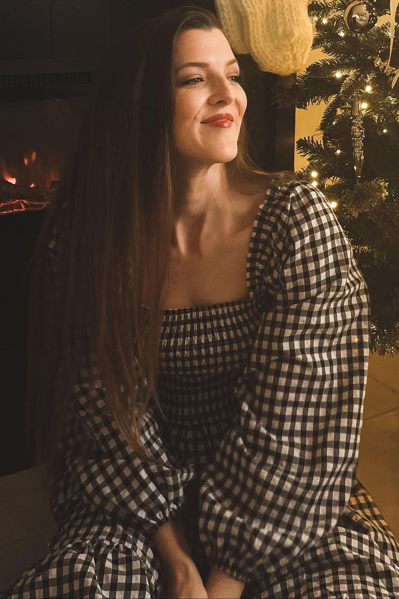 woman wearing a black and white checkered puff sleeve dress, sitting in front of a decorated Christmas tree
