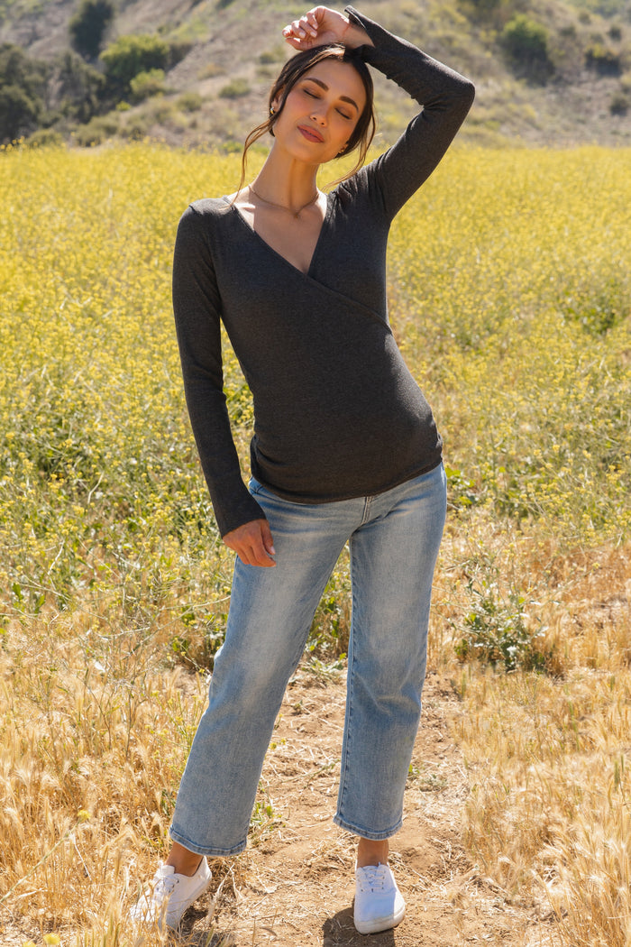 woman wearing a dark gray long sleeve maternity top with a crossover design and light blue jeans, standing in a field
