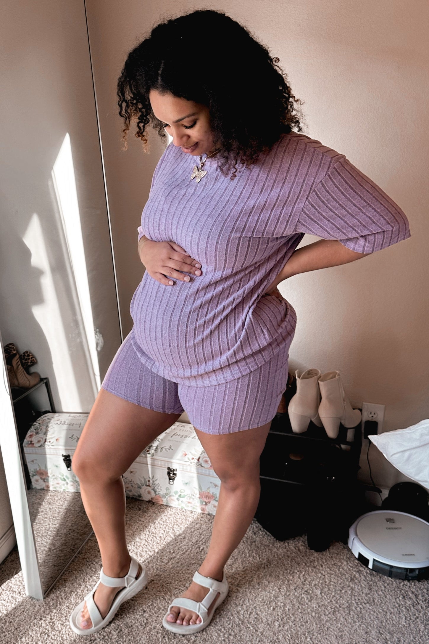 woman wearing a lilac maternity shorts set with ribbed texture, holding her baby bump and standing in a bright room