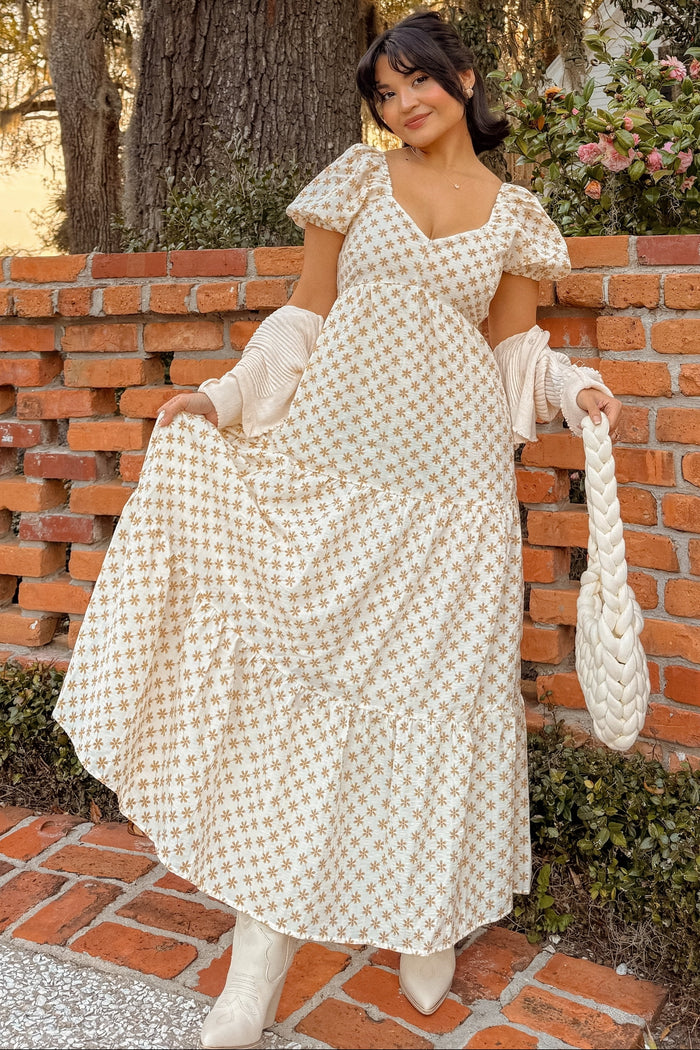 woman wearing a cream floral print maxi dress with puff sleeves and a tiered skirt, holding a braided handbag