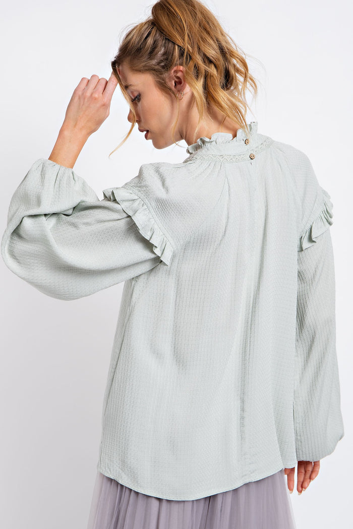 woman wearing a light green long sleeve blouse with ruffled detailing and a button closure at the back