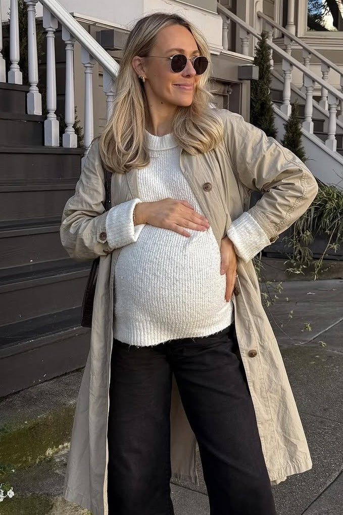 woman wearing a beige trench coat, white knit maternity top, and black pants, showing her baby bump