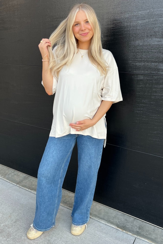 woman wearing a cream oversized maternity top and blue wide-leg jeans, holding her baby bump