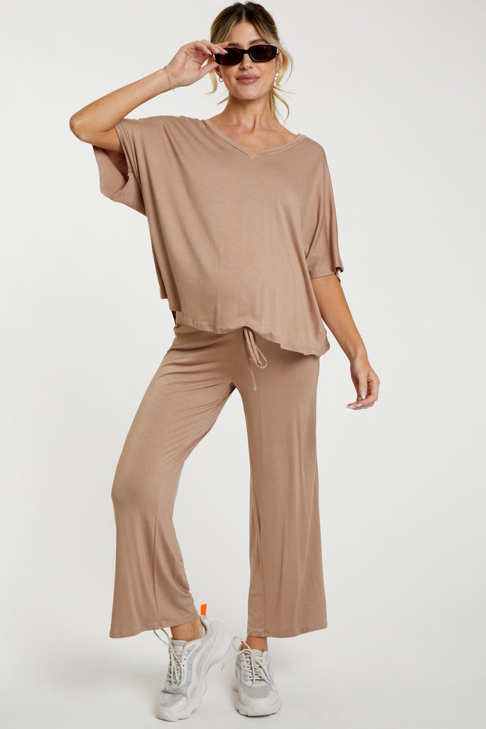 woman wearing a taupe maternity lounge set with a v-neck top and wide-leg pants, holding sunglasses and showing her baby bump