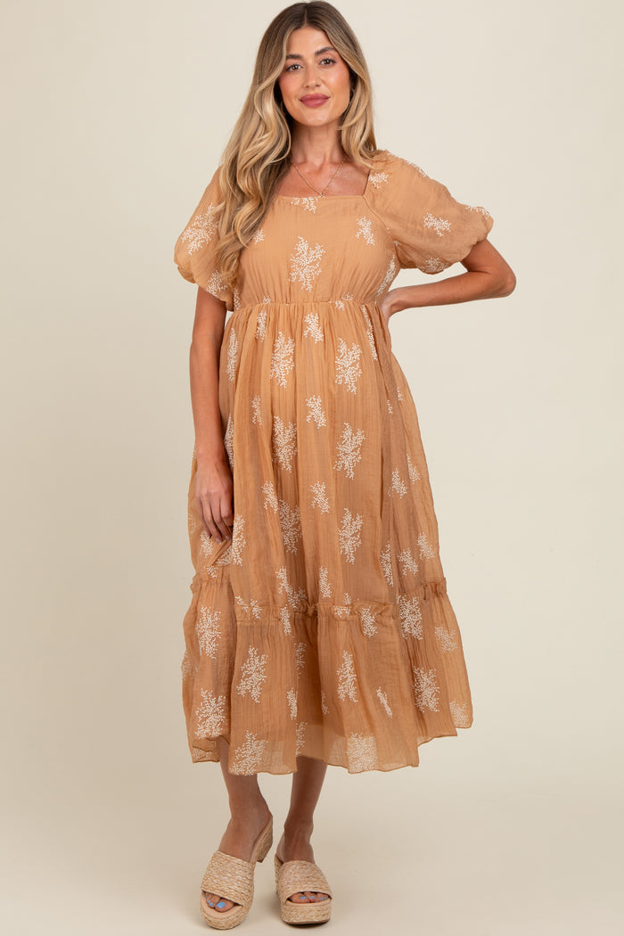 woman wearing a tan maternity dress with floral embroidery and short puff sleeves, standing and showing off her baby bump