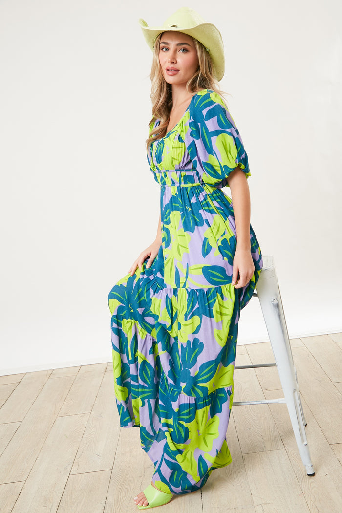 woman wearing a colorful floral maxi dress with puff sleeves and a wide-brimmed hat, sitting on a stool