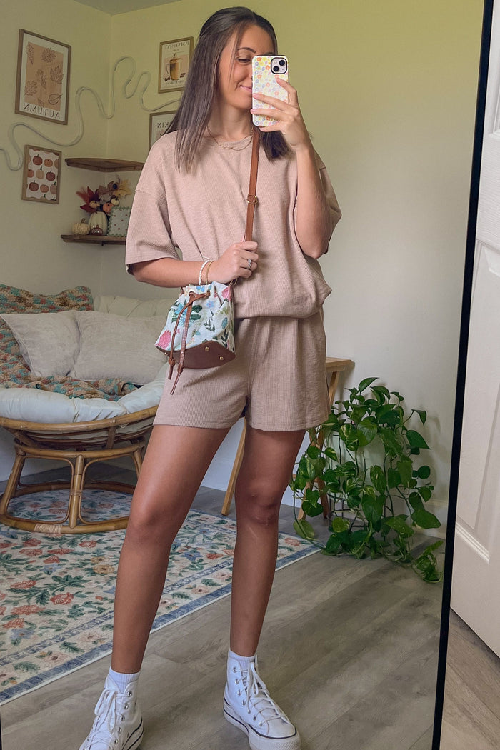 woman wearing a beige oversized short sleeve top and matching shorts, holding a floral handbag, standing in front of a mirror