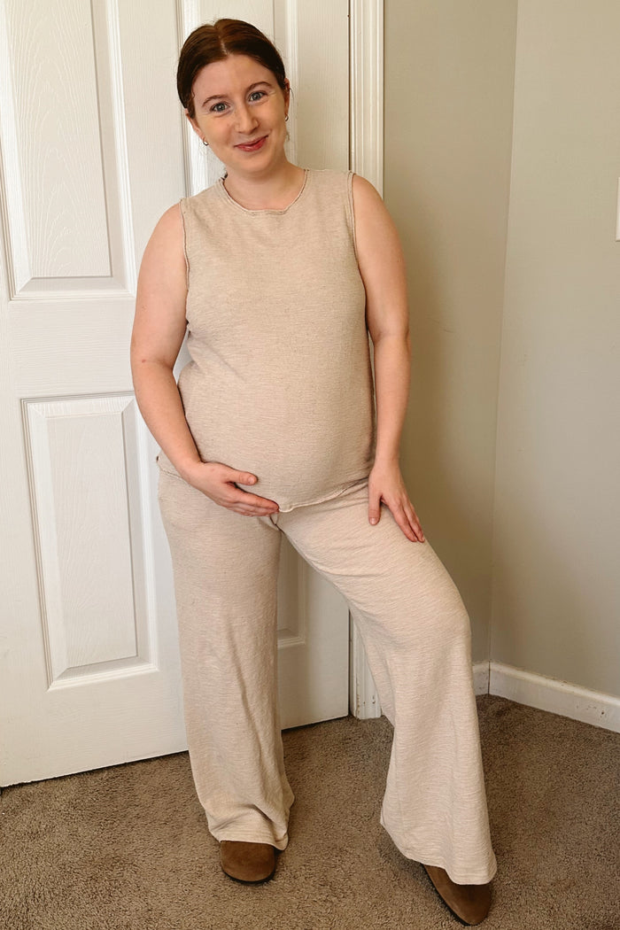 woman wearing a beige sleeveless maternity top and wide-leg pants, smiling and cradling her baby bump