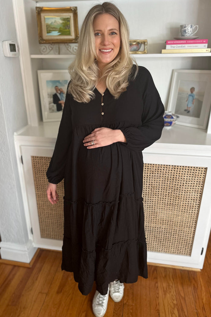 woman wearing a black maternity dress with a tiered design and button details, holding her baby bump