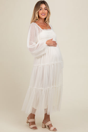woman wearing a flowing white maternity dress with a ruffled hem and long sleeves, holding her baby bump