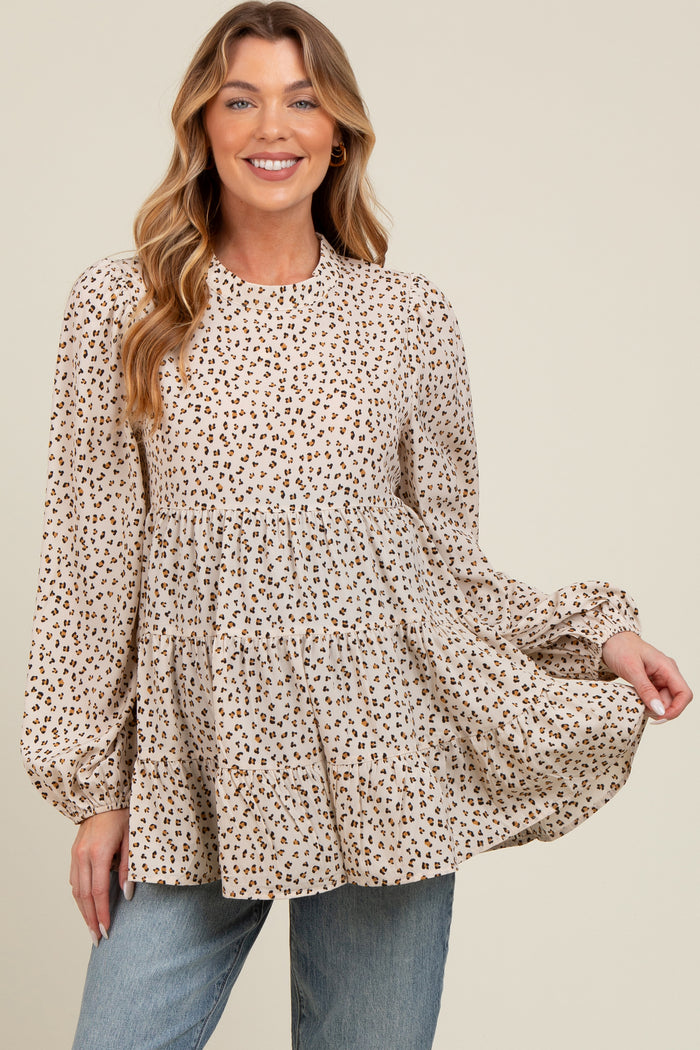 woman wearing a beige maternity top with a leopard print and tiered silhouette, smiling and holding the fabric out