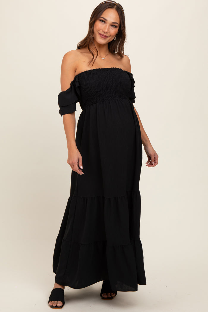 woman wearing a black off-the-shoulder maternity maxi dress with ruffled sleeves and a tiered skirt, showing her baby bump