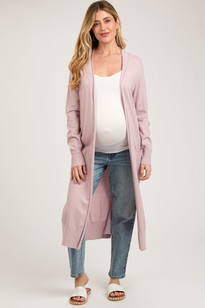 woman wearing a light pink long cardigan over a white tank top and blue jeans, showing her baby bump
