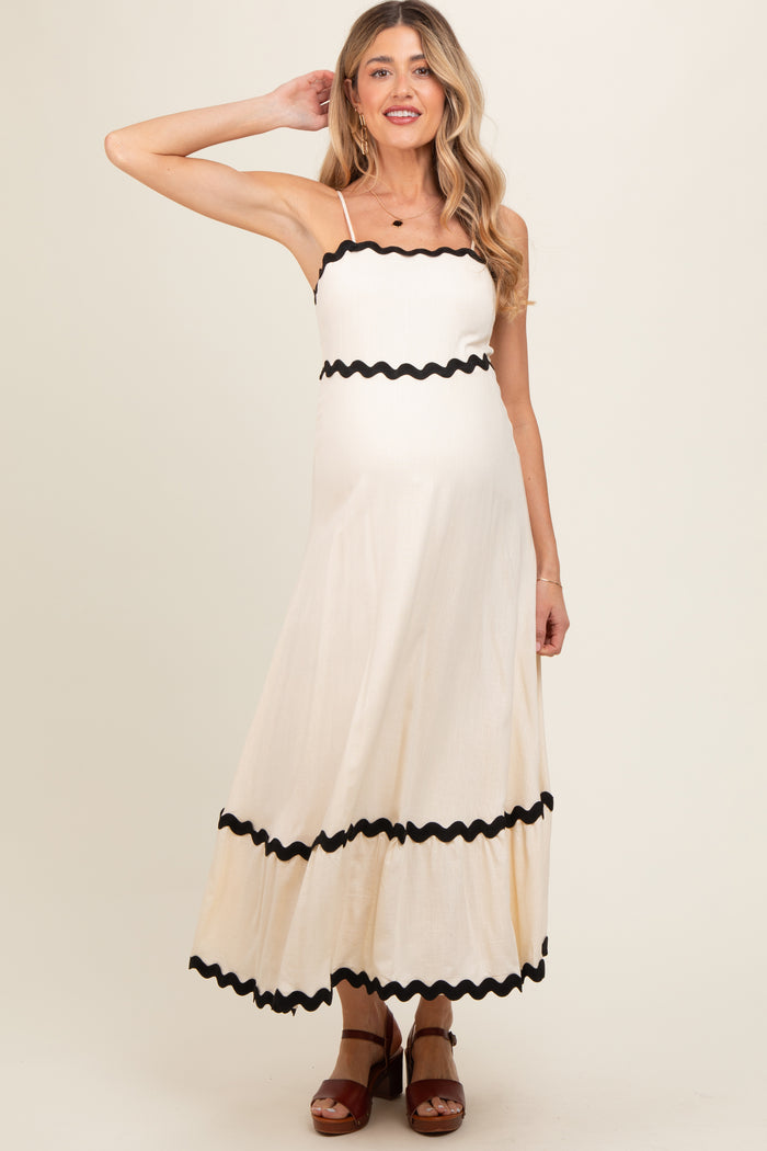 woman wearing a cream maternity maxi dress with black scalloped trim and spaghetti straps, showcasing her baby bump