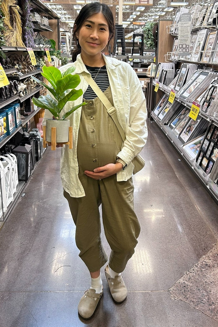 woman wearing a light green maternity jumpsuit and holding a potted plant while showing her baby bump in a store