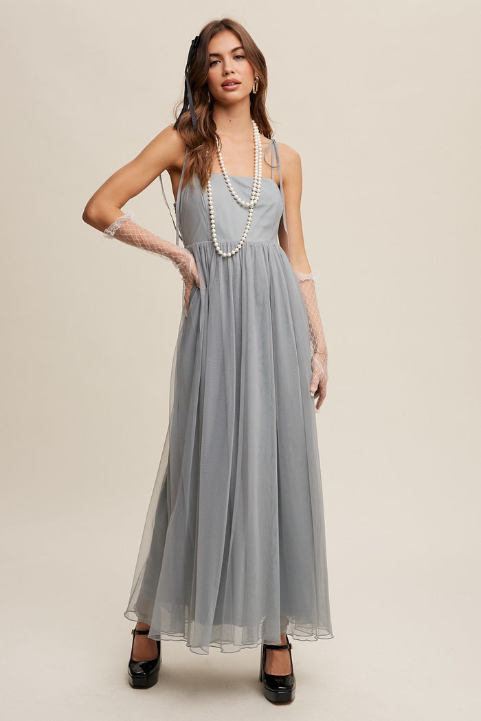 woman wearing a light gray spaghetti strap maxi dress with tulle overlay and pearl accessories, posing with gloves