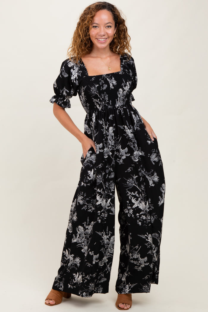 woman wearing a black floral print jumpsuit with puff sleeves and pockets, smiling at the camera