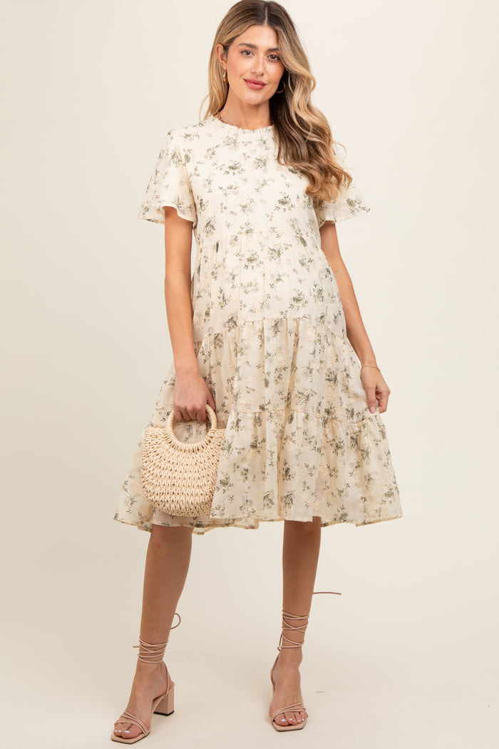 woman wearing a beige floral maternity dress with short sleeves and a tiered skirt, holding a woven bag
