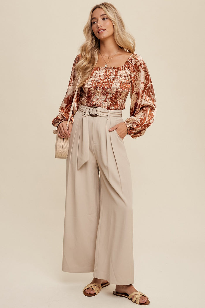 woman wearing a brown floral blouse with puff sleeves and beige high-waisted wide-leg trousers, holding a light bag