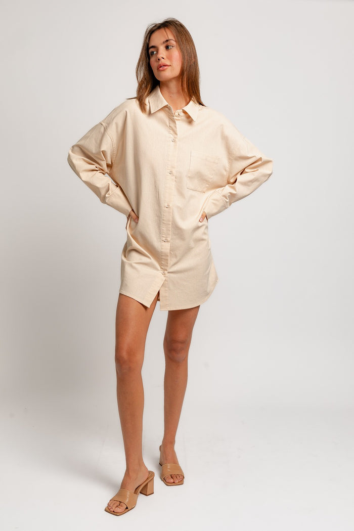 woman wearing a light beige oversized shirt dress with a pocket and button-up front, styled with nude heeled sandals