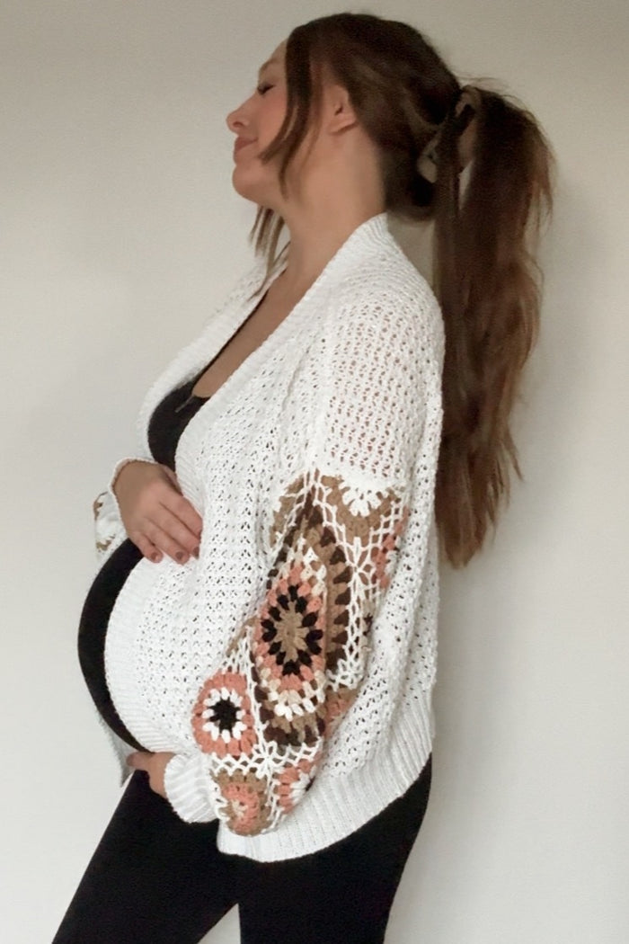 woman wearing a white open-knit maternity cardigan with colorful patterned sleeves and holding her baby bump