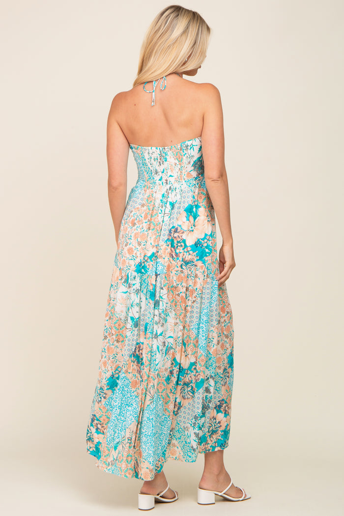Back view of a woman wearing a teal and peach floral halter maxi dress with white block heel sandals