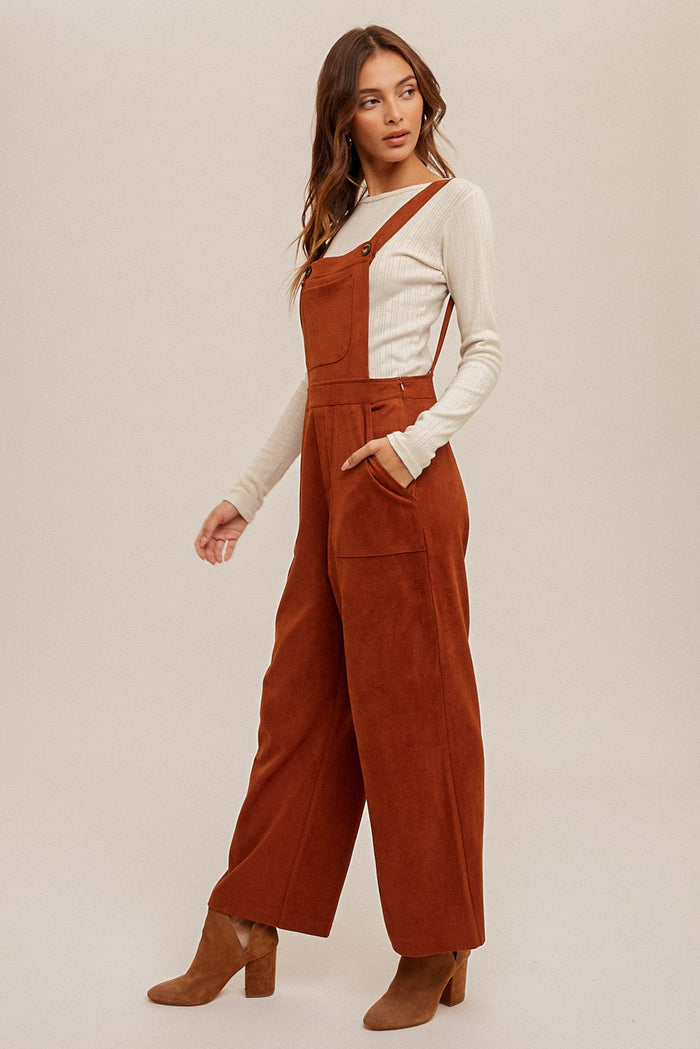 woman wearing rust-colored corduroy overalls with a white long sleeve shirt underneath and tan heeled ankle boots